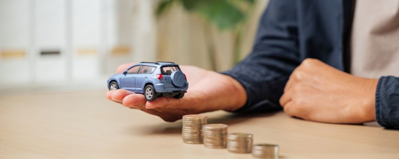 Closeup of a man hand holding a blue toy car model with ascending stacks of coins nearby highlights auto finance car loan payments.