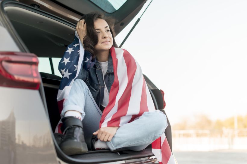 A young woman holds a large US flag in the trunk of her car.