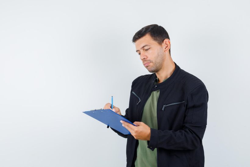 Young male taking notes on clipboard in t-shirt, jacket and looking busy.
