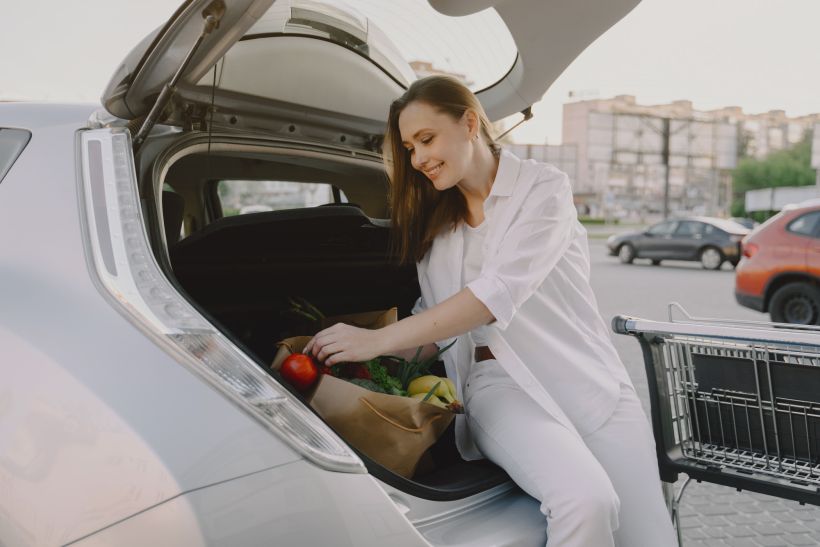 A smiling woman sits in the trunk of her car with groceries.