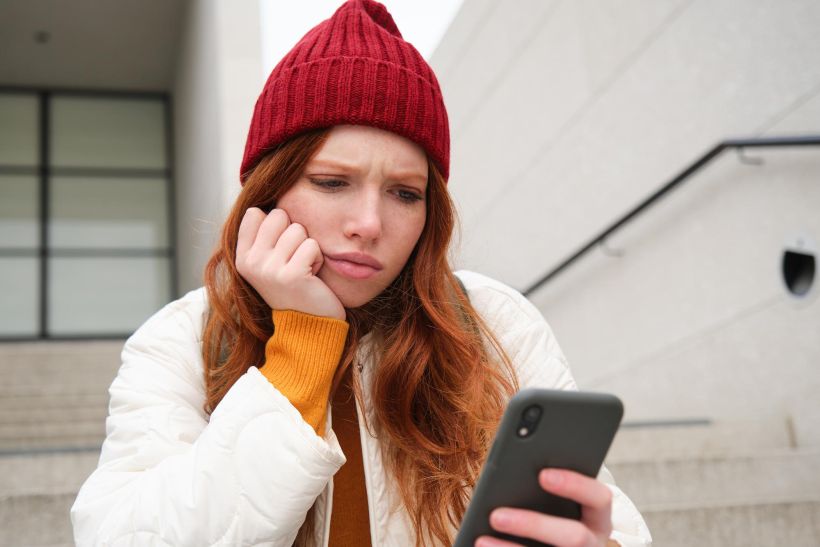 Portrait of sad redhead girl looks upset and disappointed at smartphone screen.