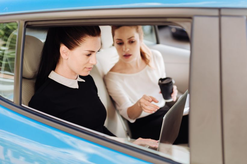 Nice pleasant businesswoman sitting in the car with her colleague and pointing at the laptop screen.