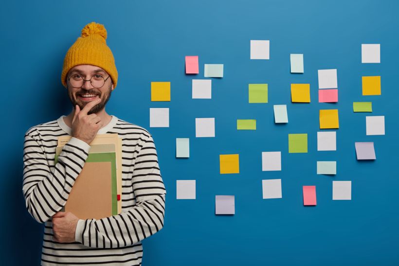 Cheerful male student holds chin stands with notebook and textbook uses sticky notes on blue wall.