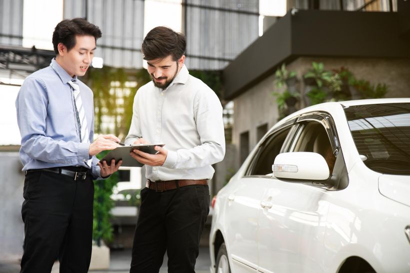 A young man signs documents while standing next to a car.