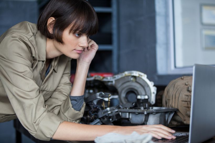 A female mechanic leans on a table and uses a laptop.
