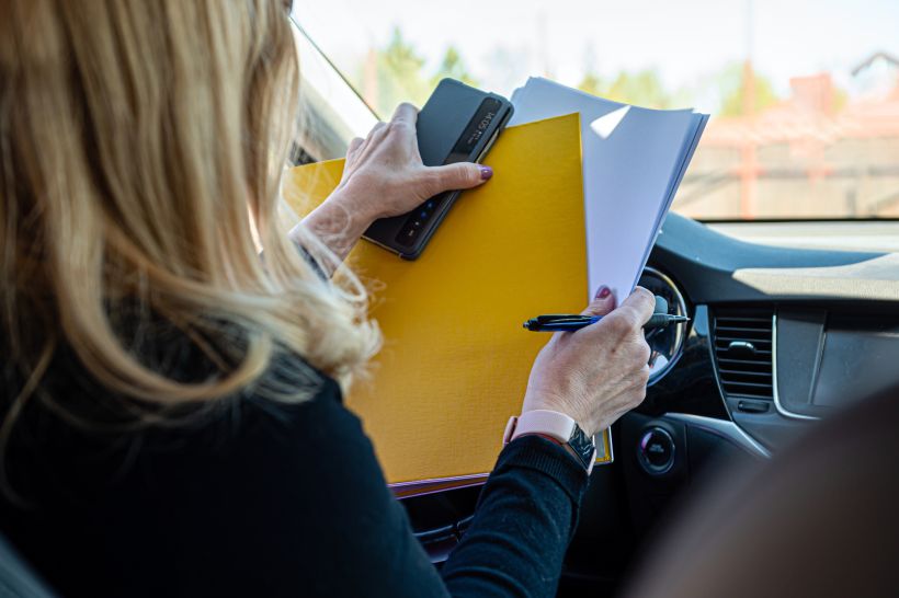 Middle aged blonde business woman in glasses in car working with documents rear view person.